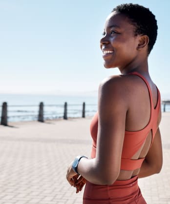 A smiling woman in athleisure standing outside near a body of water as she goes for a walk as part of her mental health routine.