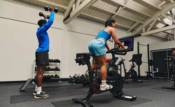 Two friends doing strength and cardio workouts side by side in a gym using a Peloton Bike.