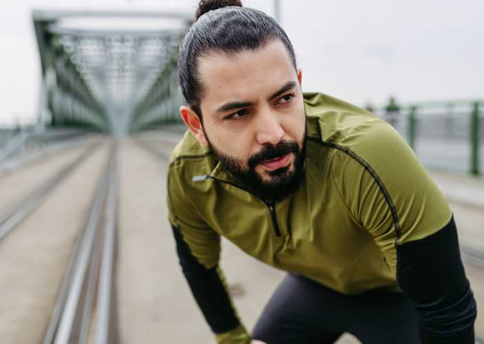 A man exhaling as he puts his hands on his knees while standing up outside. He's dealing with a stomach ache after exercise.