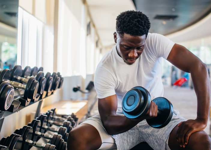 A man sitting at the gym curling a dumbbell with one arm while sitting on a workout bench.