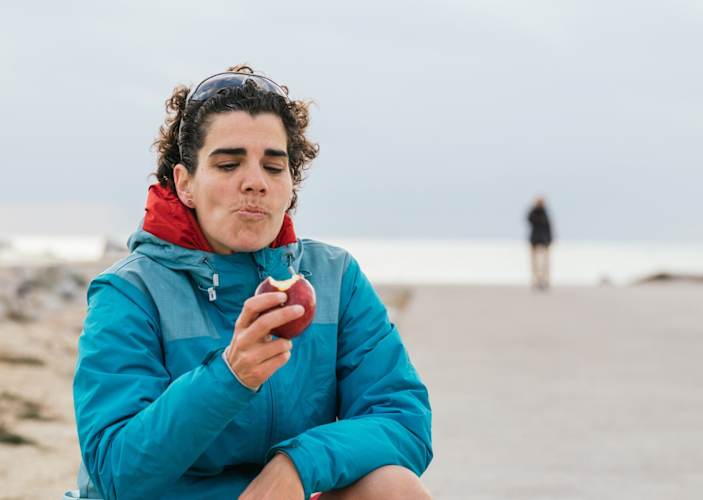 A runner eating an apple shortly before, during, or after a run on the beach. She is preventing runger by taking in some fuel.