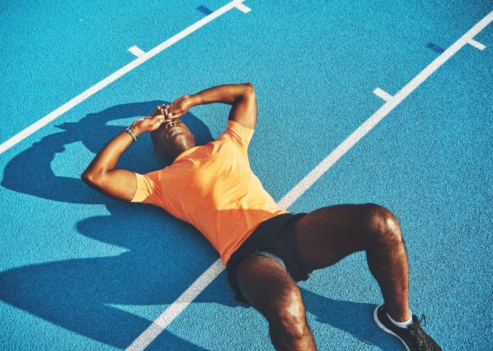 A male athlete experiencing bonking. He is exhausted, lying down on his back with his hands on his head, on a blue track.