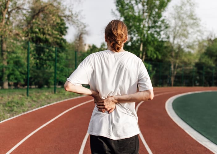 A photo of the back of an athlete who is on an outdoor track. They are feeling bloated after a workout and holding their back with their hands. 