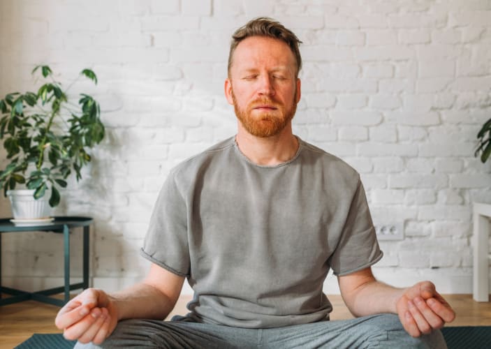 A man practicing a meditation for stress at home in a bright, sunny room. His eyes are closed and his hands are resting on his knees as he sits in a cross-legged position.