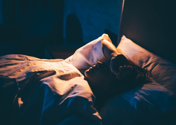 A woman lying in bed wide awake at night with her arm on her head in distress. She's dealing with circadian misalignment.