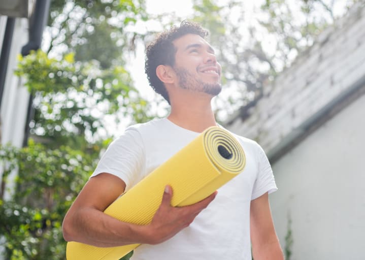A man holding a yellow yoga mat and smiling while standing outside. Learn how to motivate yourself to work out here.
