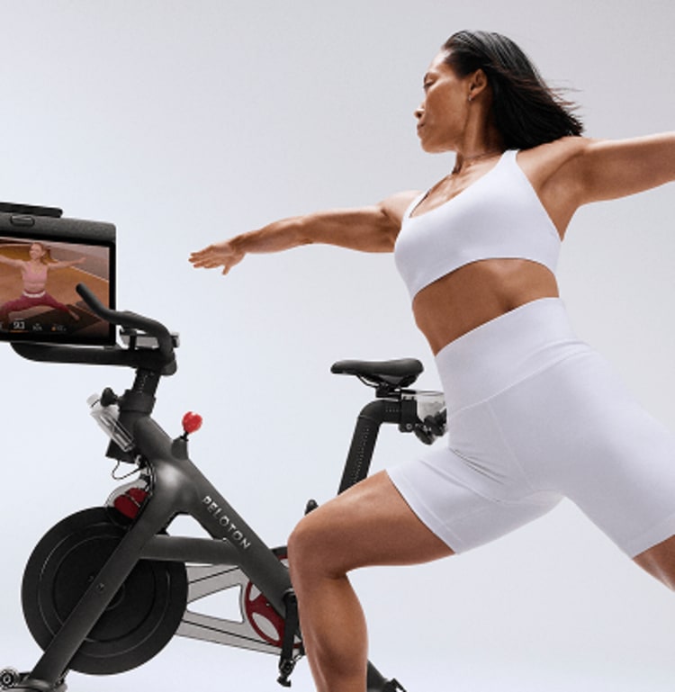 A lady performs stretch beside a Peloton bike whilst following a Peloton class on the Peloton bike swivel screen
