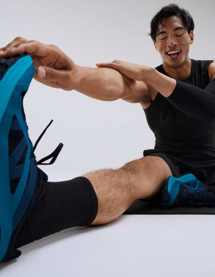 Man smiling sat on a grey floor with a Peloton Cross Training Bike behind him whilst he is sat on the floor one leg in a cross leg position the other leg is stretched out in front where the man is doing a calf stretch by holding the toe of his sneakers