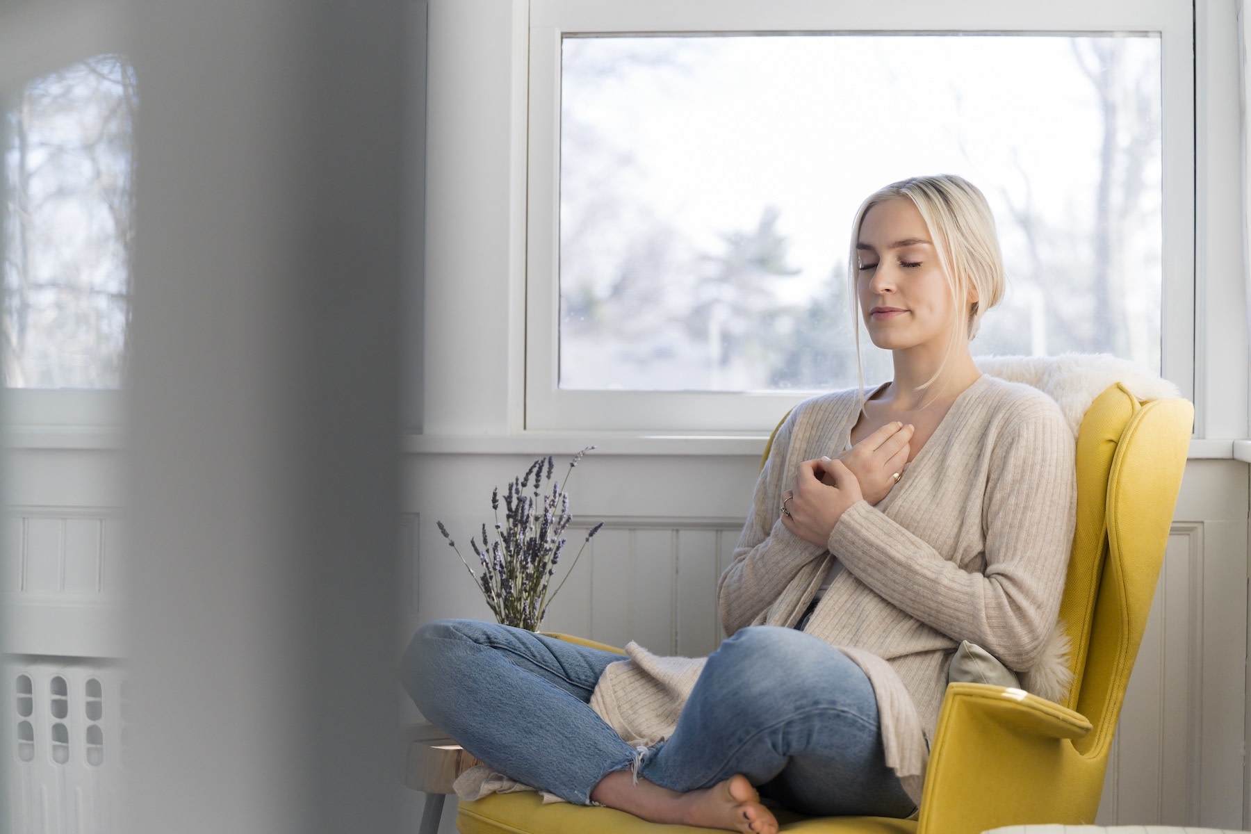 A woman sitting in a chair at home and meditating. Her hands are on her chest, her eyes are closed, and her legs are in a criss-cross position. Learn eight meditation tips in this article.