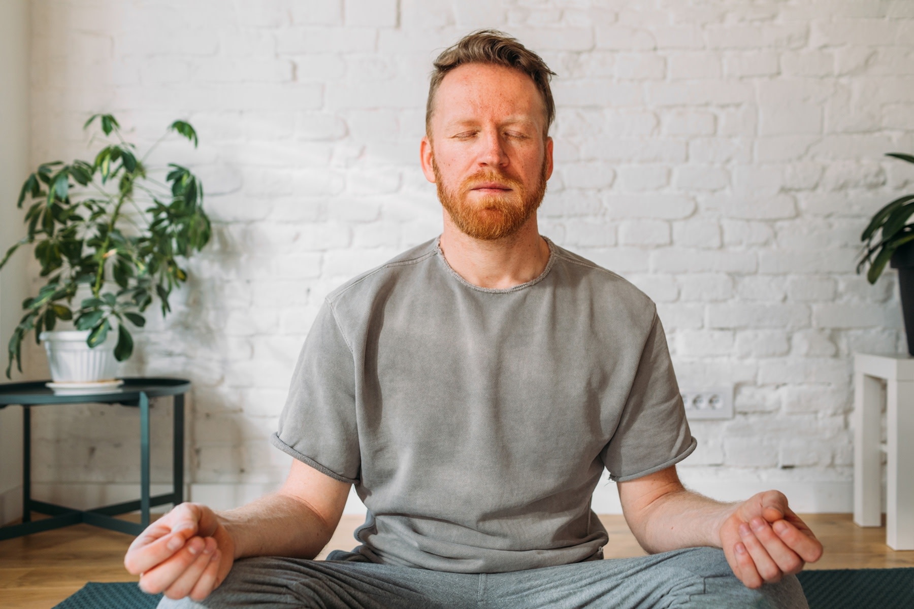 A man practicing a meditation for stress at home in a bright, sunny room. His eyes are closed and his hands are resting on his knees as he sits in a cross-legged position.