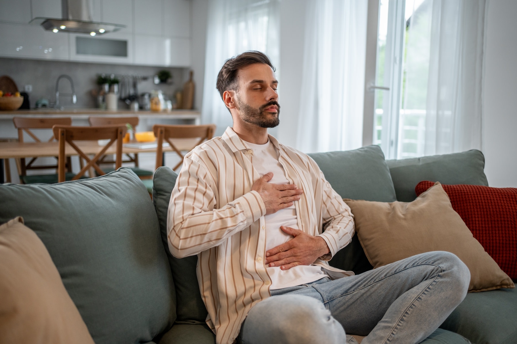 A man sitting on a cough at home practicing belly breathing. His eyes are closed and he has one hand on his chest and the other on his belly.