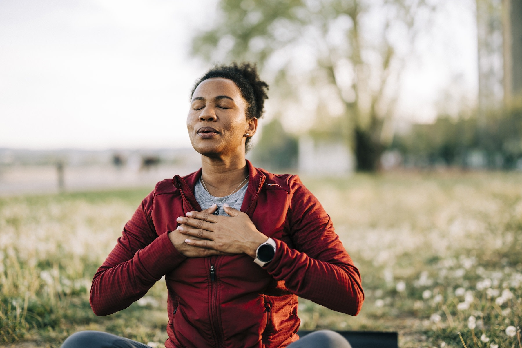 A woman calmly sitting down and practicing cyclic sighing. Her hands on her chest and she's exhaling through her mouth as she sits outside.