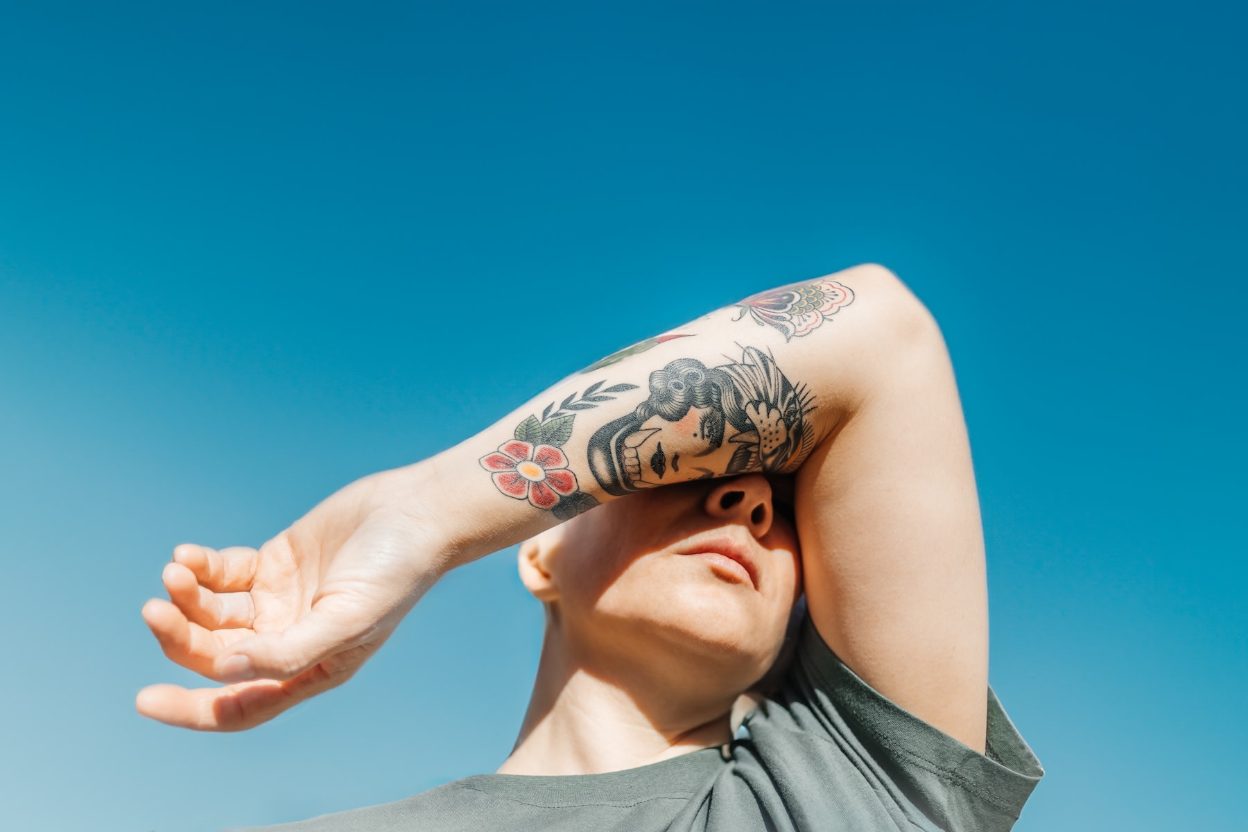 A person covering their eyes with their tattooed arm. They are feeling sick from stress. The background is a bright blue sky.