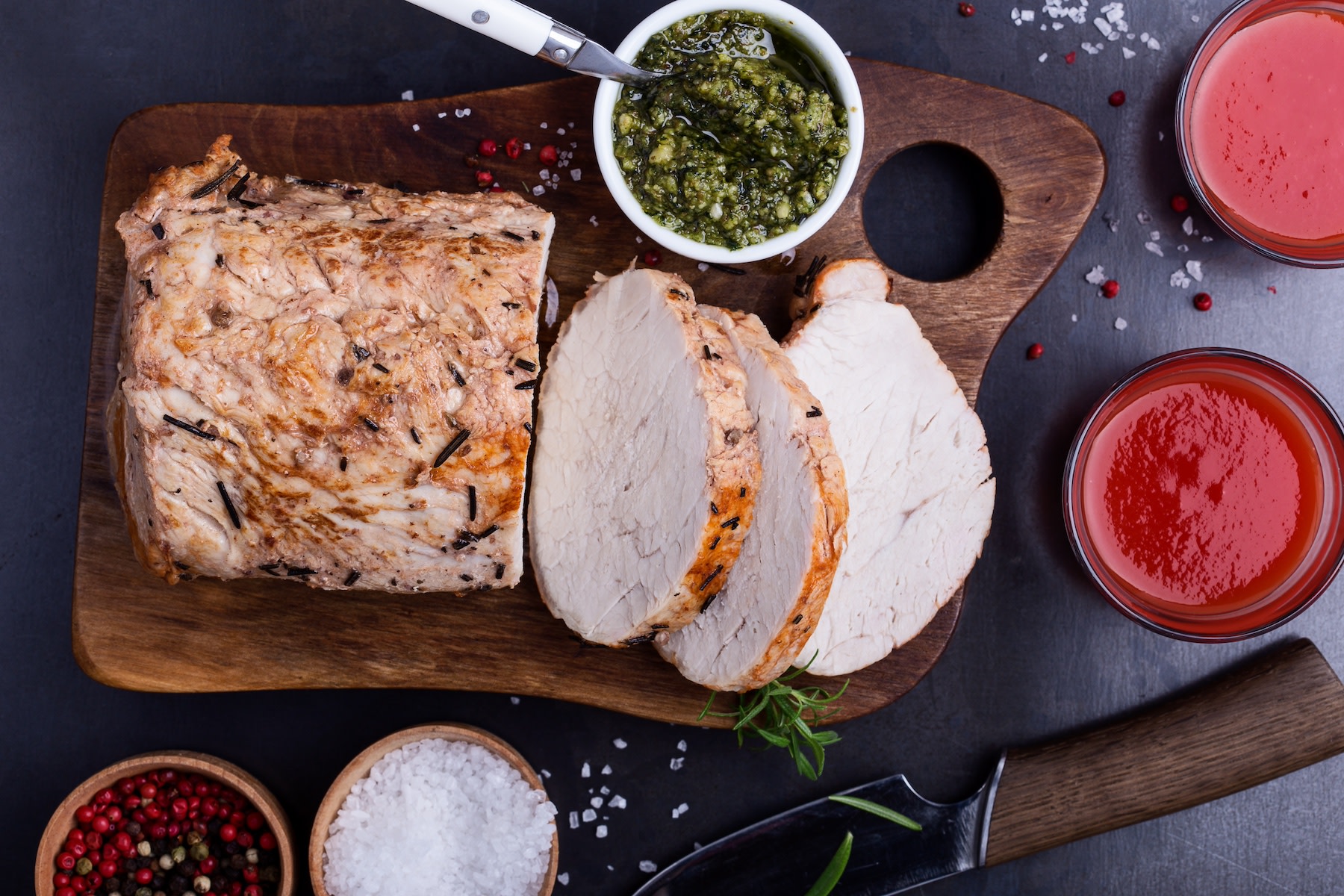 Herb-roasted pork loin, a lean protein food, resting on a cutting board next to pesto sauce.