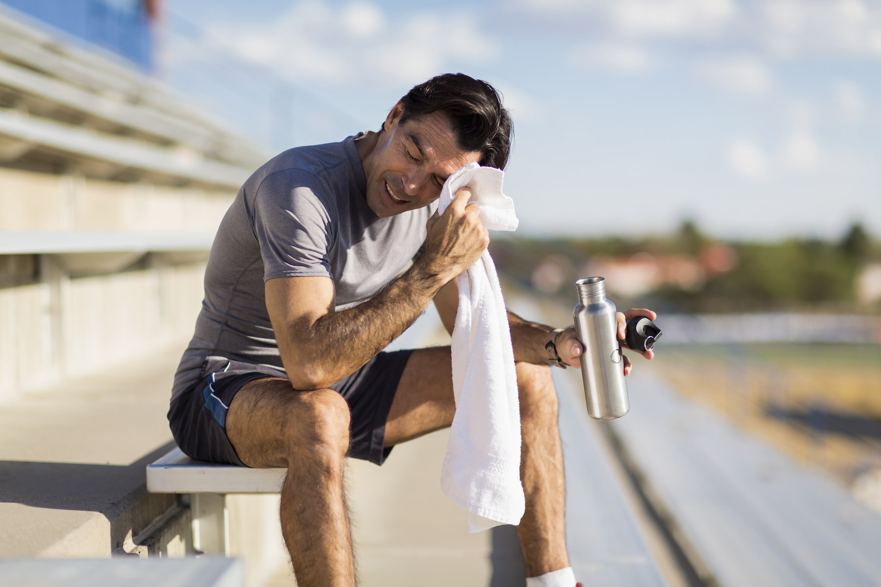 Why Do I Sweat So Much? - In this image, a middle-aged man is sitting on bleachers outside after a workout while smiling, holding a water bottle, and wiping away sweat from his face with a towel.