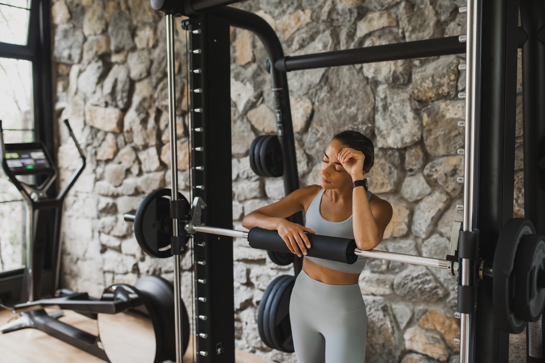 A woman in the gym near a barbell set. Her eyes are closed and her hand is on her head as she tries to work out with a headache.