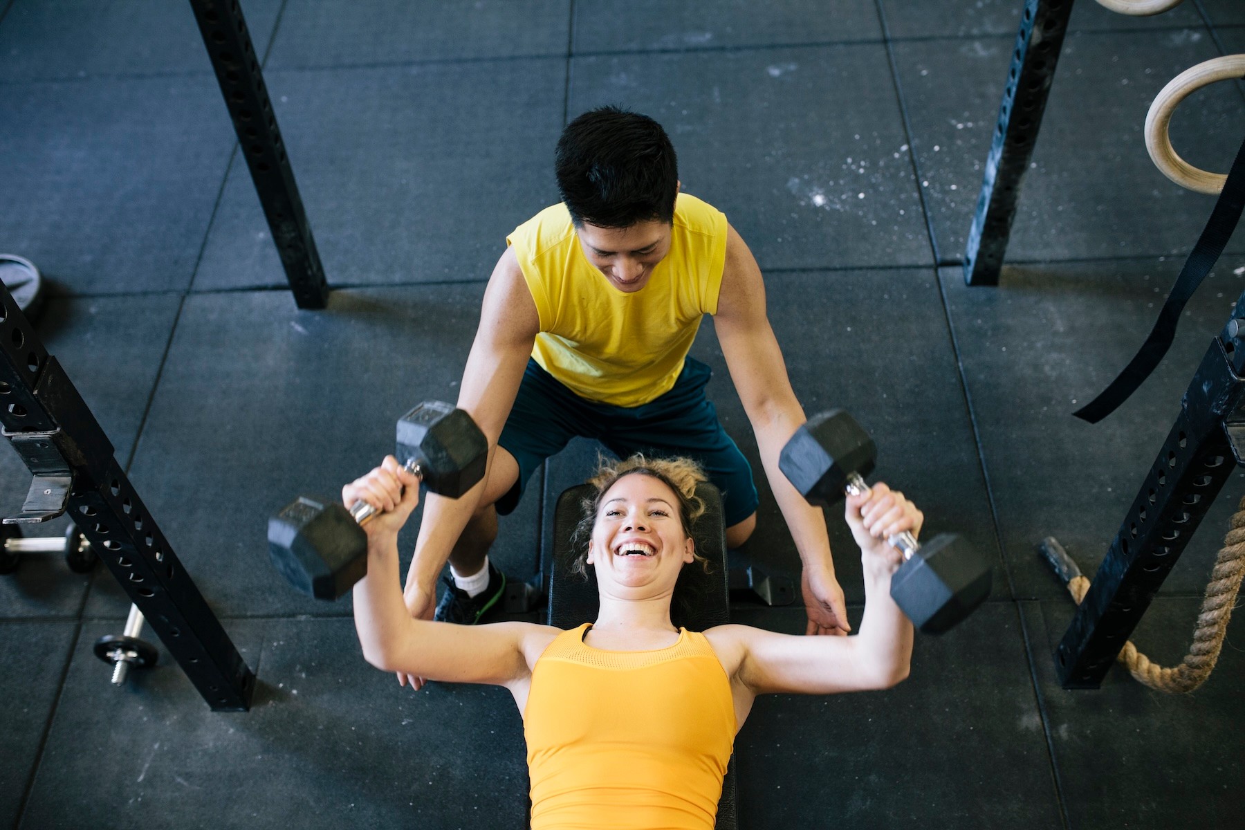 A man spotting a woman who is lifting dumbbells on a workout bench. She practiced proper gym etiquette and asked him for a spot at a convenient time.