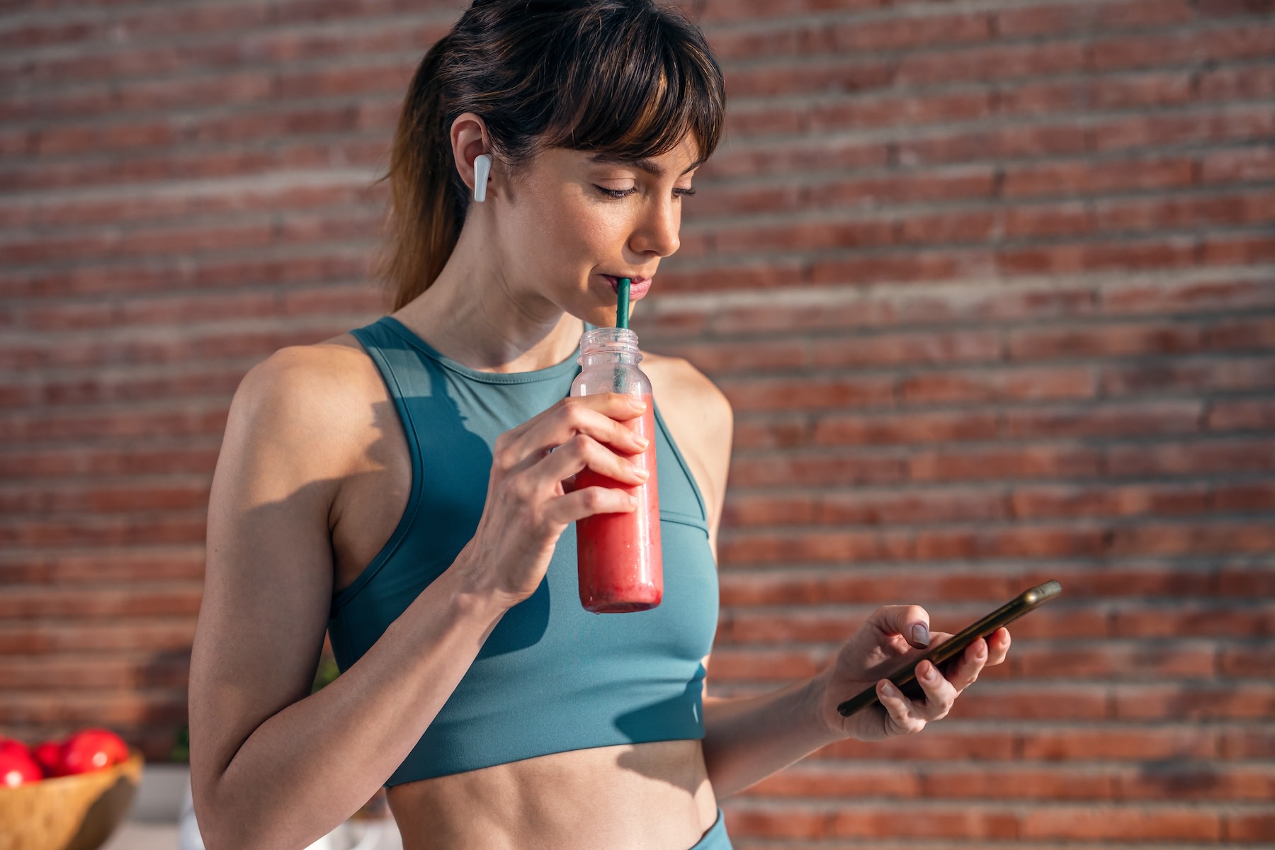 A woman drinking a glass of watermelon juice through a straw before a workout while looking at her phone.