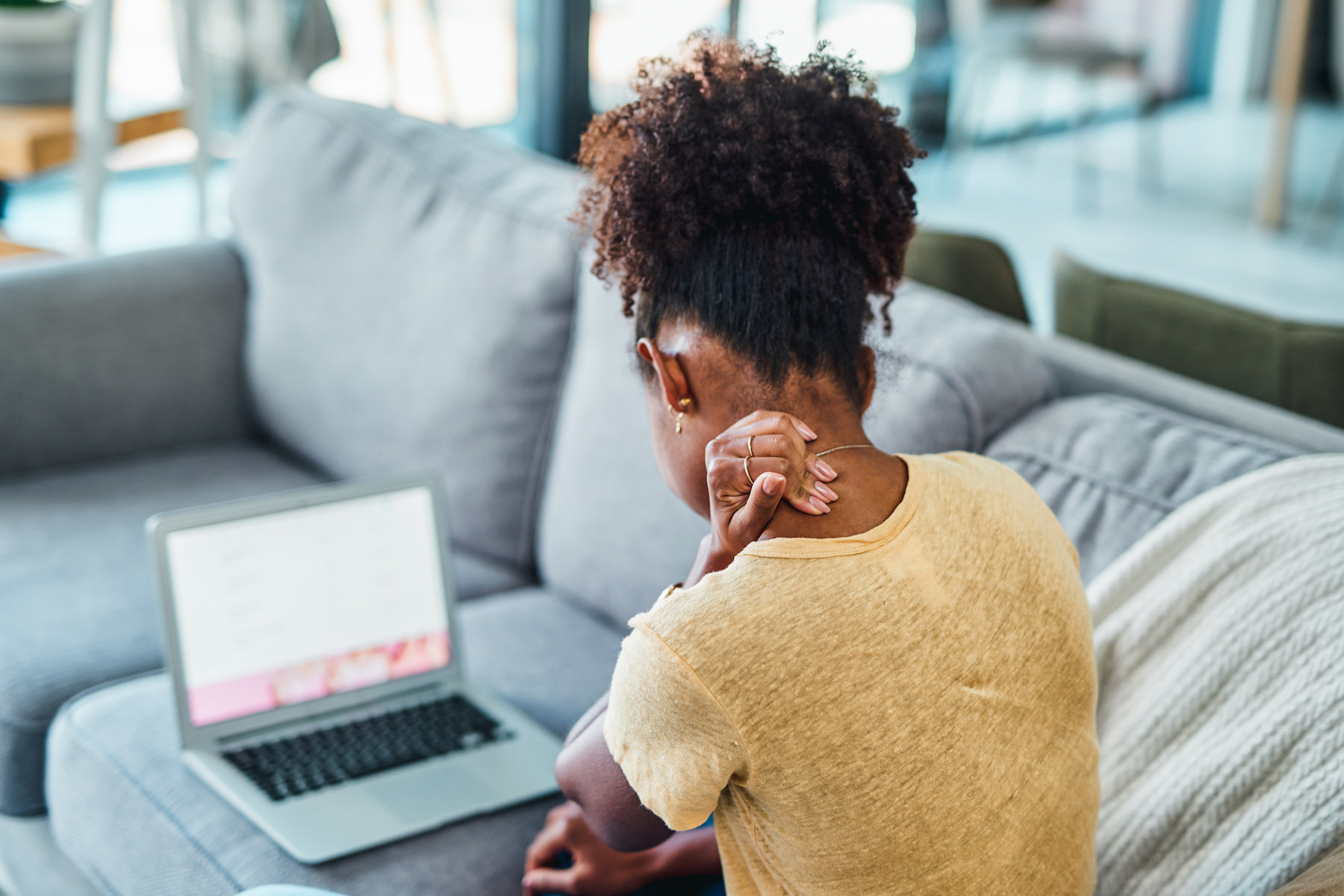 A woman experiencing tech neck. She is rubbing her neck in pain while sitting on a couch and looking down at her laptop.