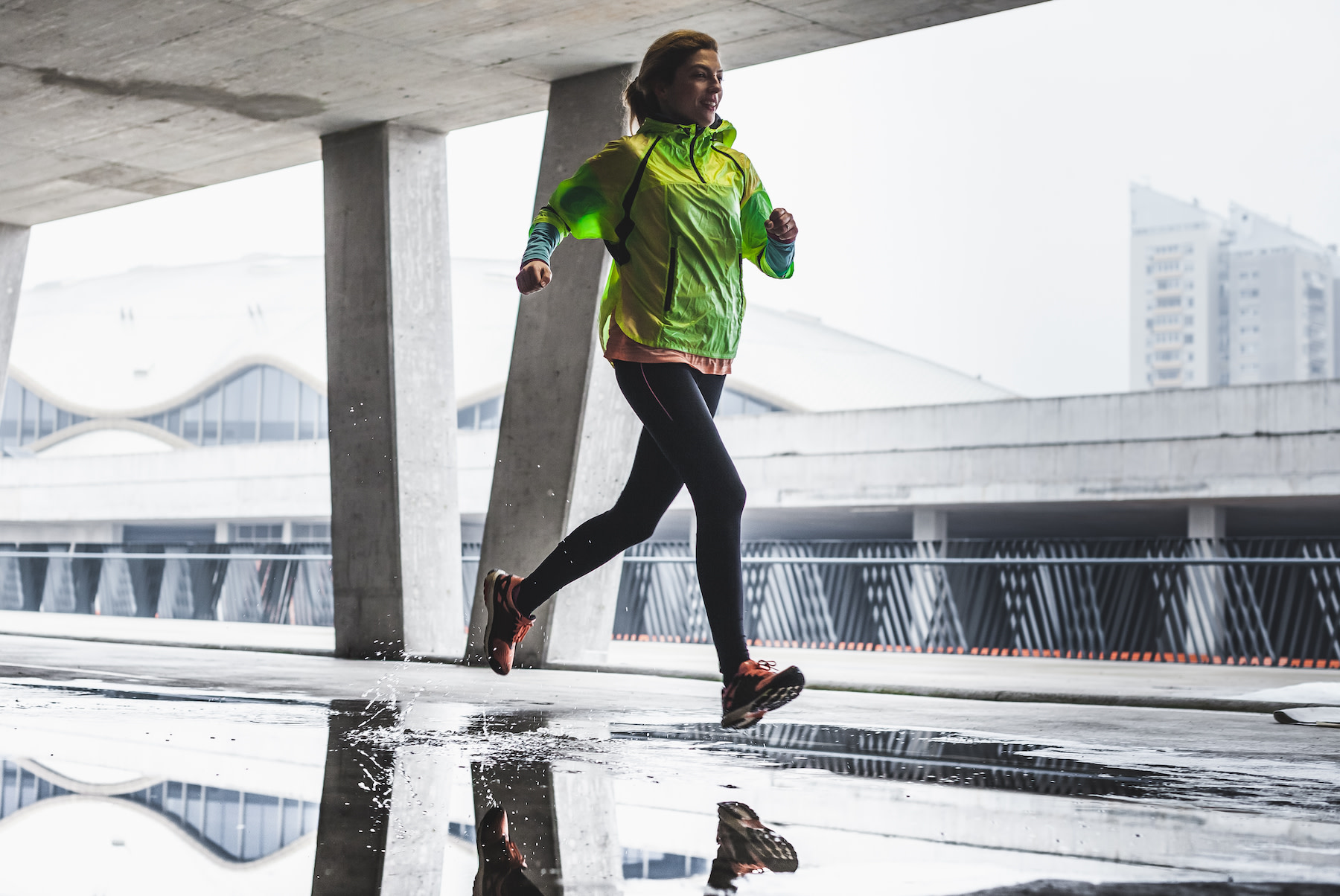 A woman smiling and running in the rain. She is wearing a bright yellow jacket with black leggings and running shoes.