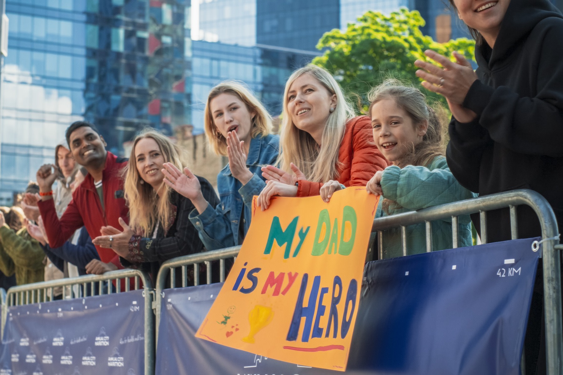 A line of spectators leaning up against a fence and cheering at a marathon. A little girl in the crowd is holding a colorful marathon sign that says "My dad is my hero."