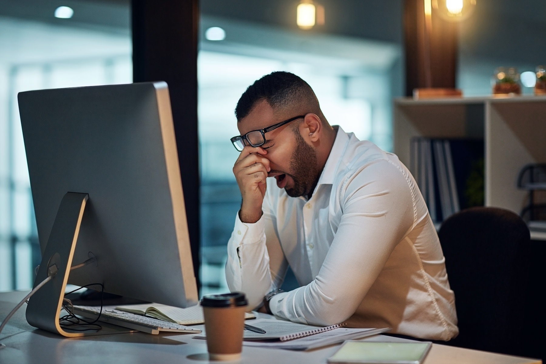 A man who only got six hours of sleep is yawning and rubbing his face while sitting at his work desk.