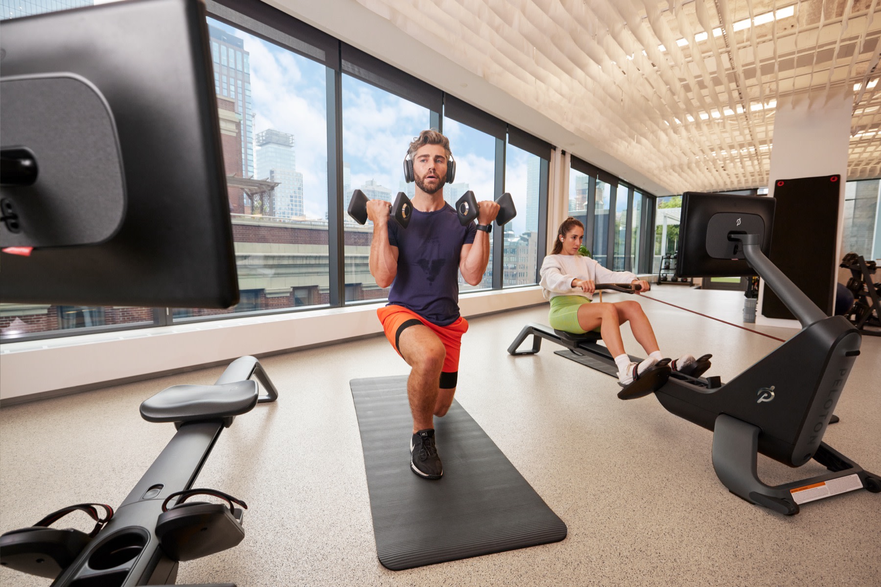 Two exercisers in fitness center, one lifting dumbbells on mat off of Peloton Row and one rowing on Peloton Row