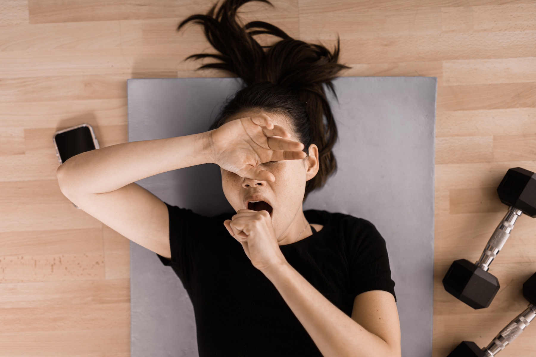 A woman yawning during a workout. She's lying down on a yoga mat next to her phone and a pair of dumbbells and covering her mouth and eyes as she yawns.