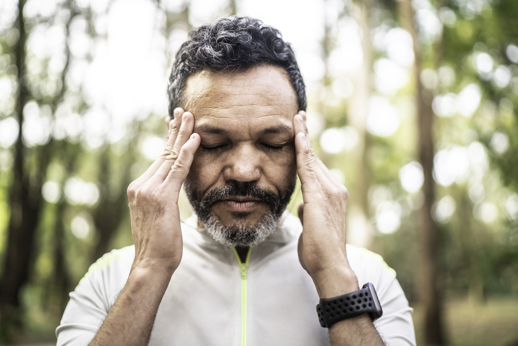 A man standing outside and holding his hands to his temples as he suffers from a headache after a workout.