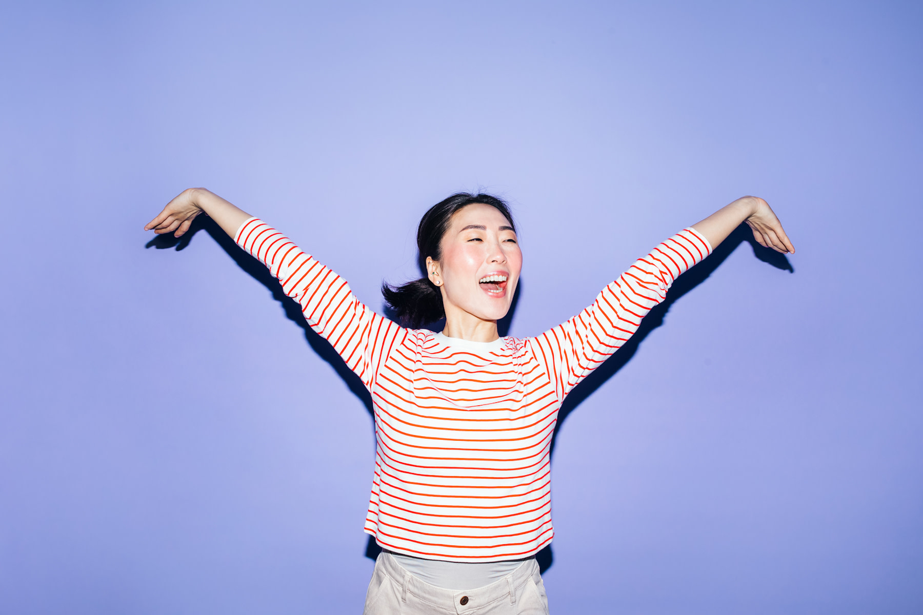 A woman smiling and spreading out her arms energetically. She's wearing a red and white striped shirt and standing against a lavender background. Learn how to get more energy in this article.