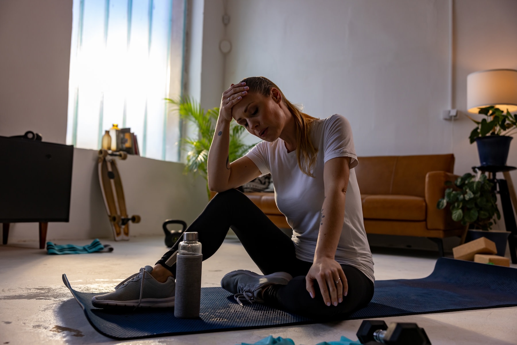 A woman sitting down on a yoga mat at home with her head resting in her hands and her eyes clothes as she takes deep breaths. She felt dizzy when stretching.