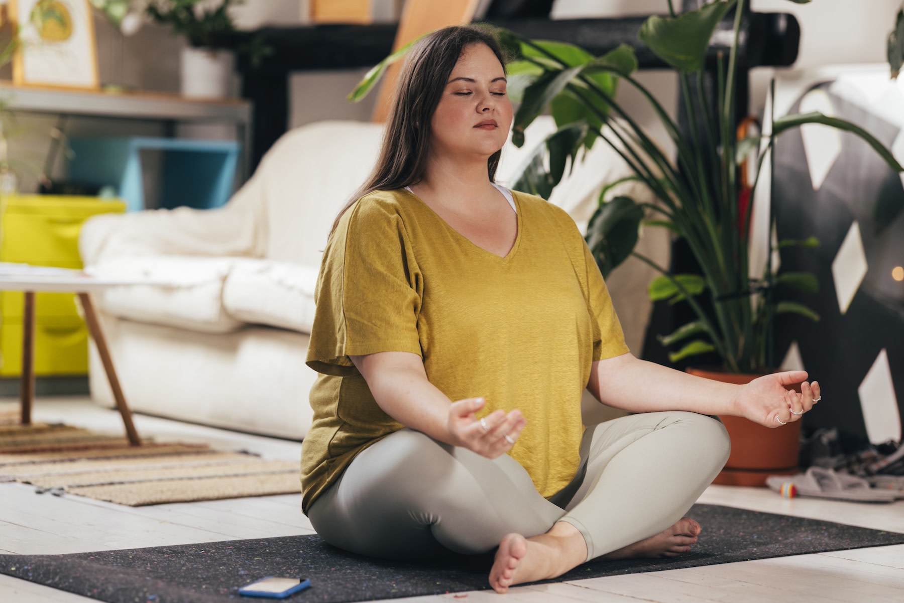 A woman meditating in lotus position, sitting with her eyes closed on a yoga mat at home. Learn about the benefits of meditating before or after a workout in this article.