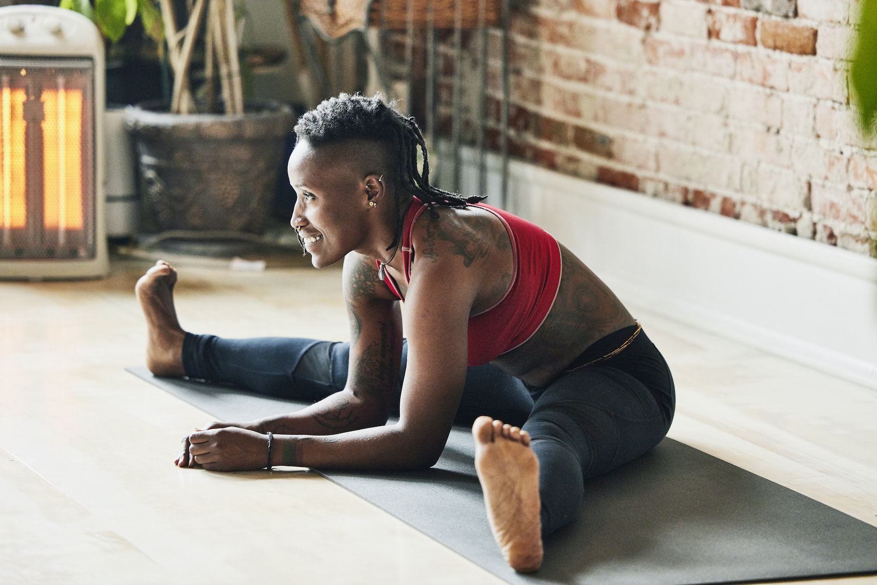 A smiling yoga instructor with tattoos sitting down and stretching on their mat in a sunny room. Learn more about whether or not you can work out after getting a tattoo in this article..