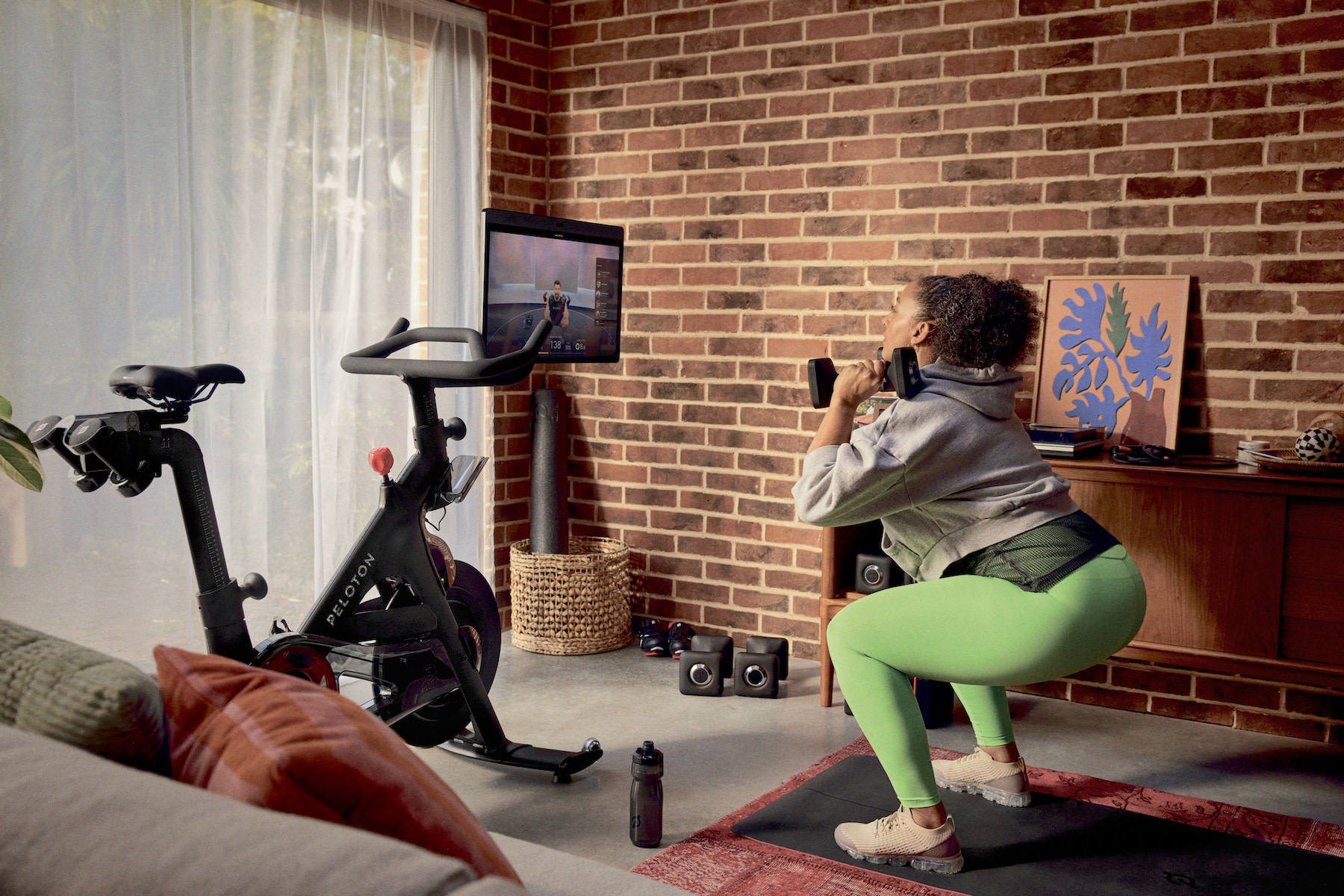 Woman does squat exercise with weights next to a Peloton Bike