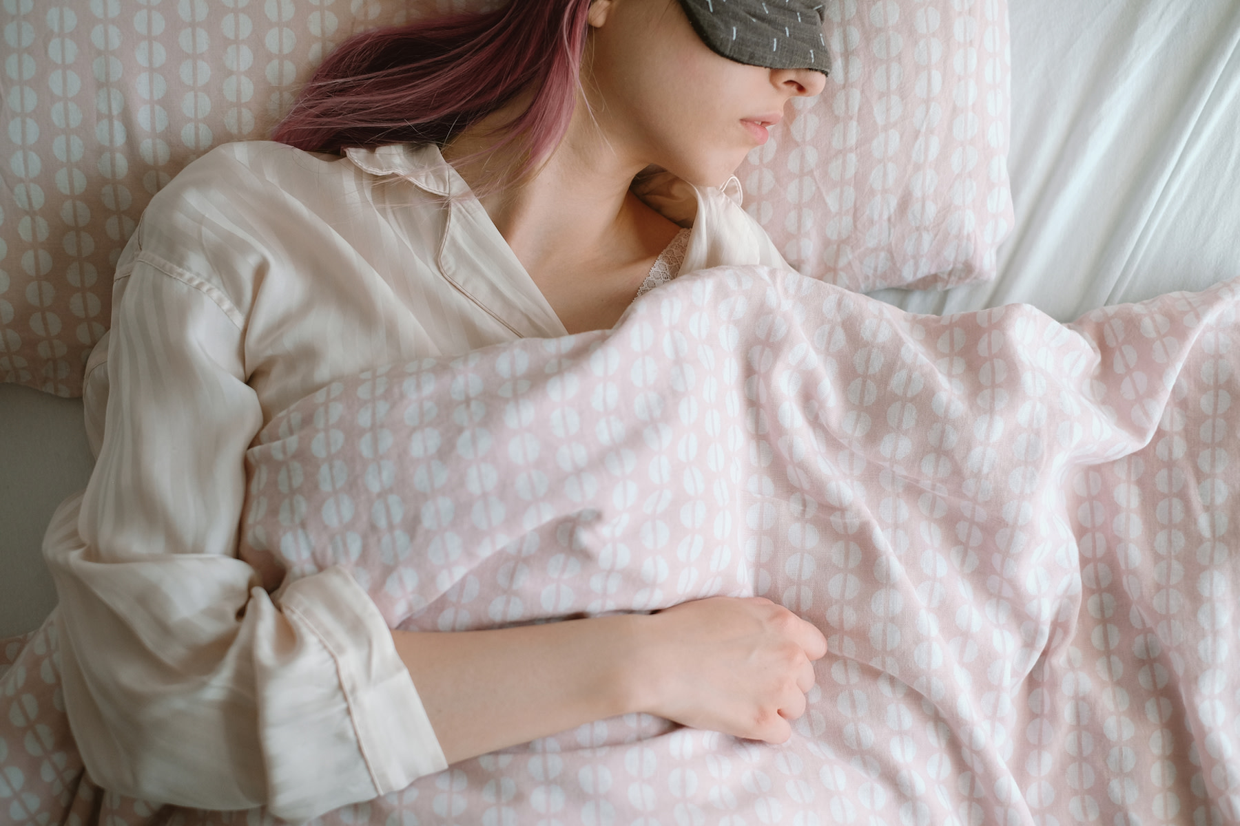 A woman with pink hair and an eye mask sleeping soundly in bed at the best temperature for sleep.
