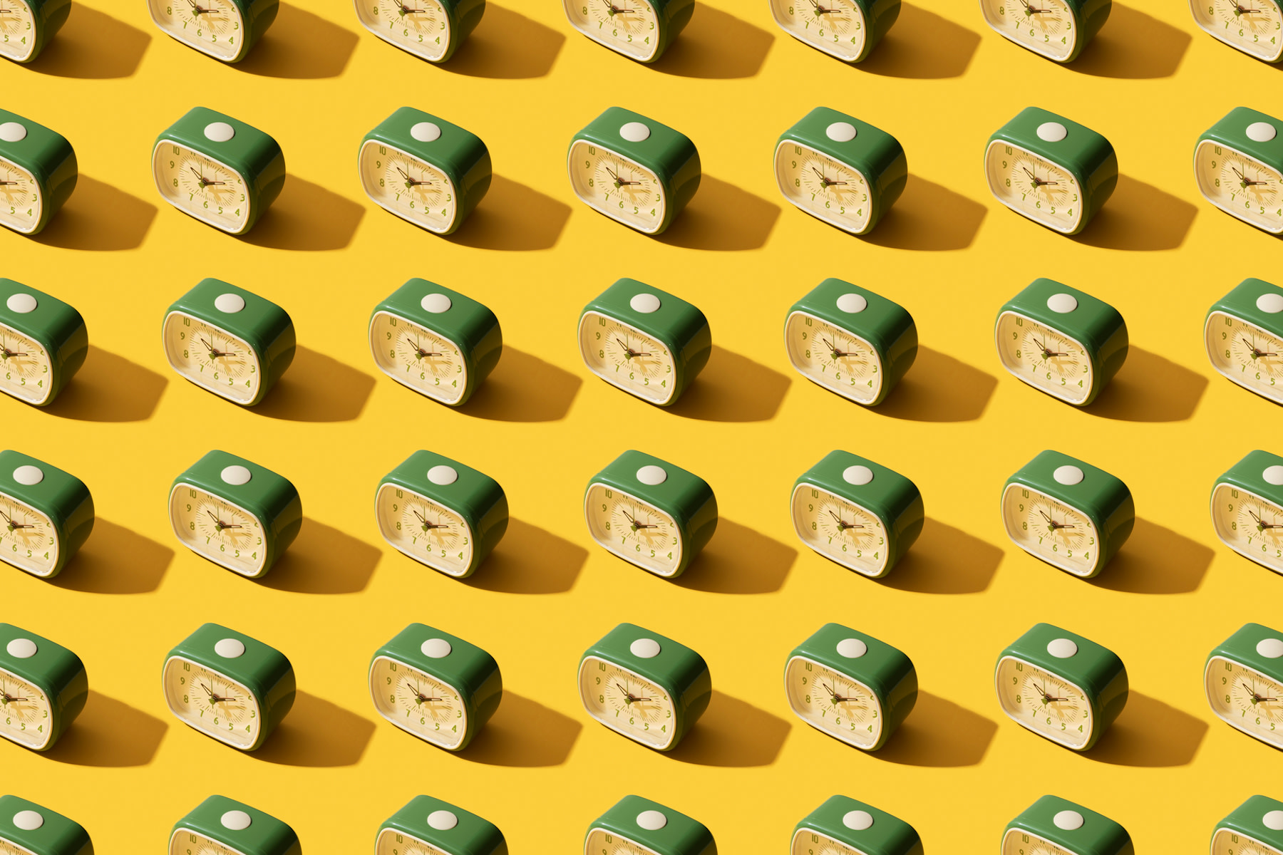 In this image for an article about how to wake up early, there are many old-school, identical green alarm clocks shown on top of a yellow background.