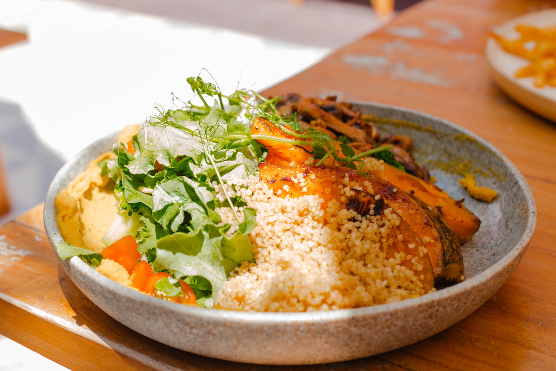 A high-protein grain bowl featuring quinoa, leafy greens, and pumpkin. The bowl is sitting on a wooden table in the sunlight.