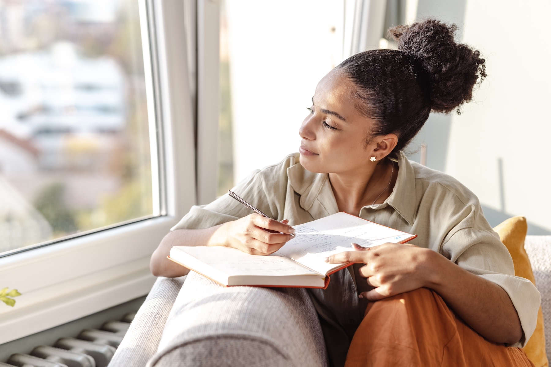 A young woman sitting on her couch and practicing a gratitude exercise by writing in her gratitude journal.