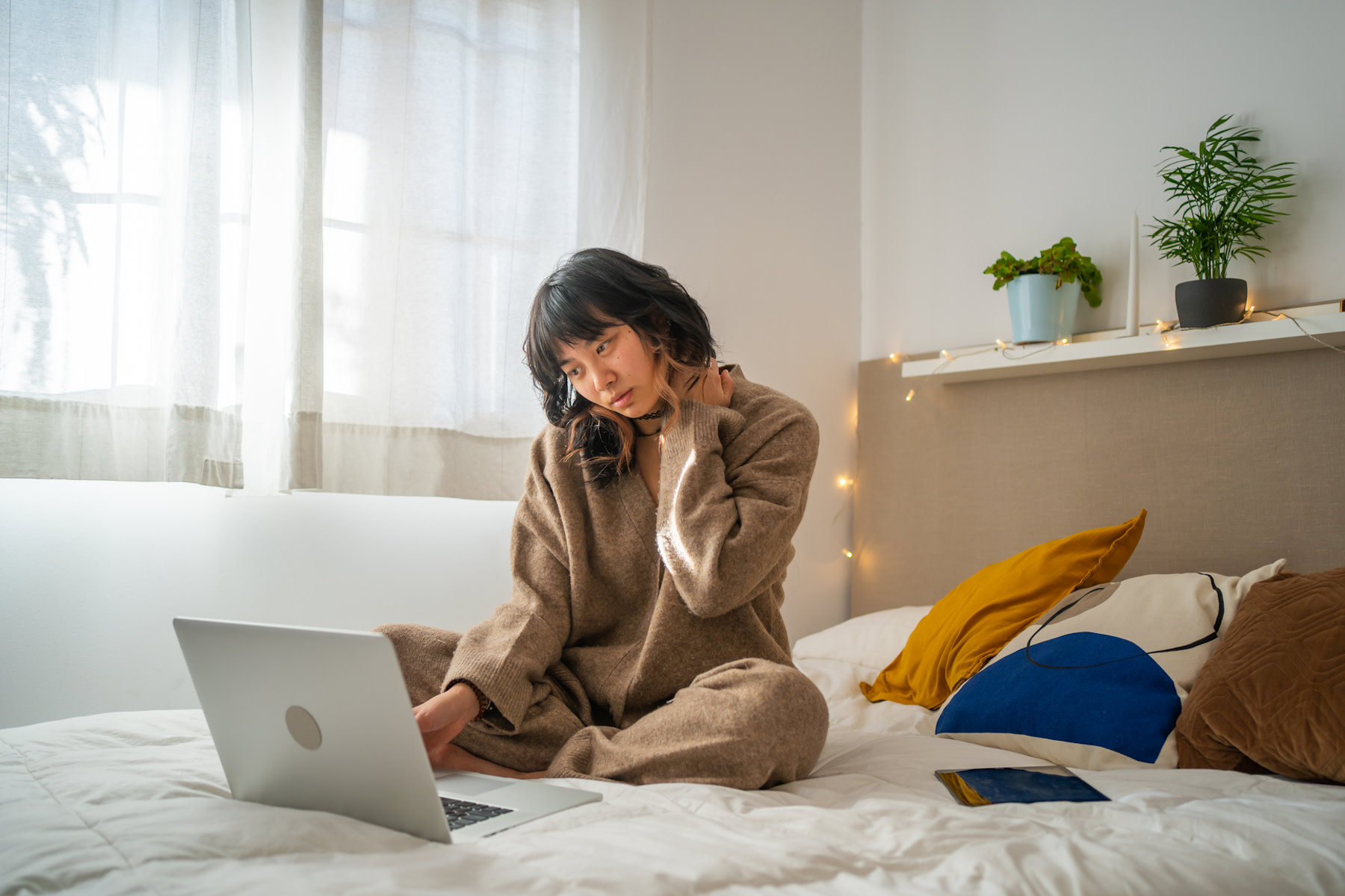 A woman experiencing tech neck. She is rubbing her neck while sitting in bed and looking down at her laptop.