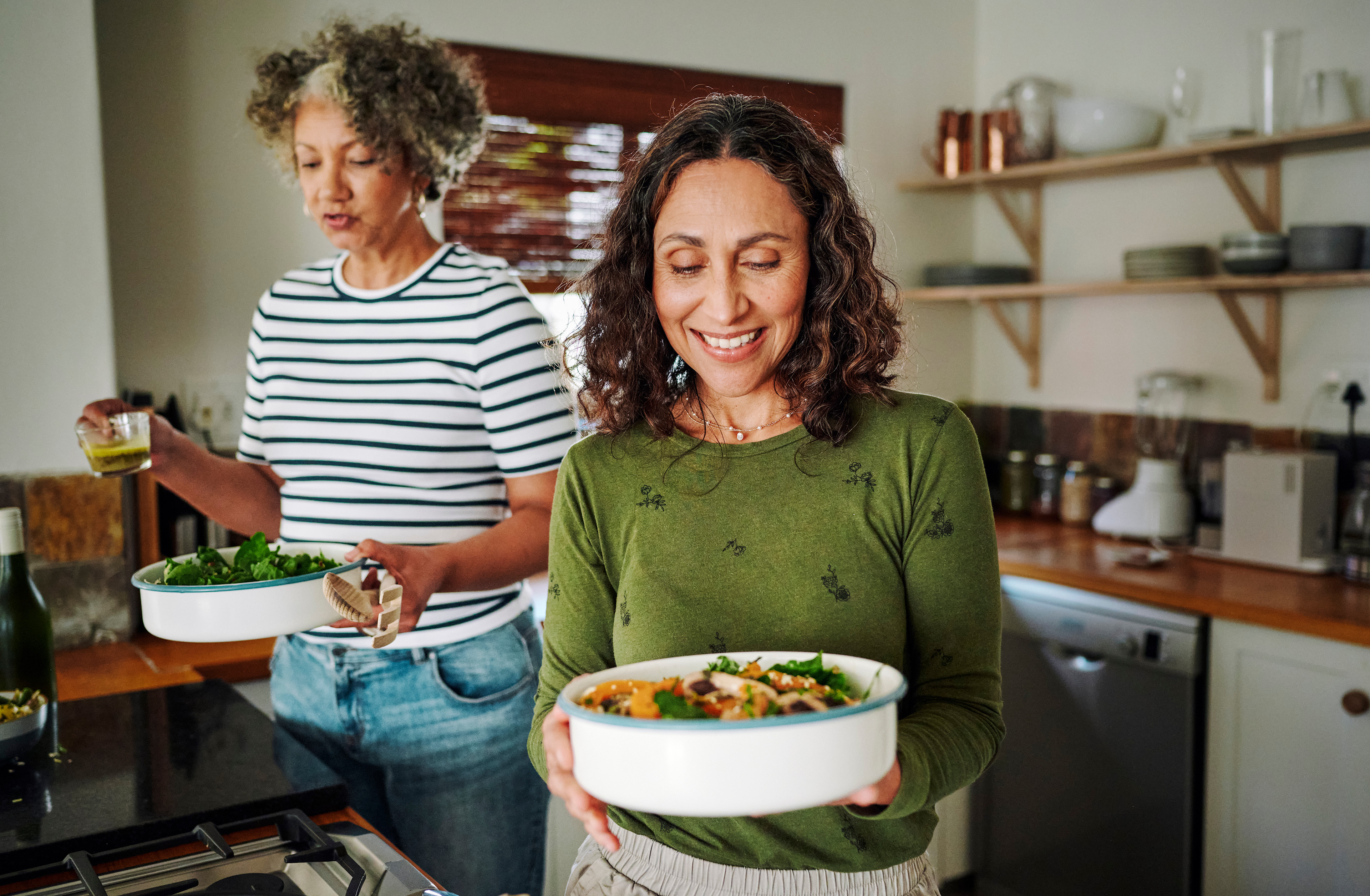 Two happy middle-aged women prioritizing menopause nutrition and preparing to eat a healthy meal with fresh veggies and other ingredients.