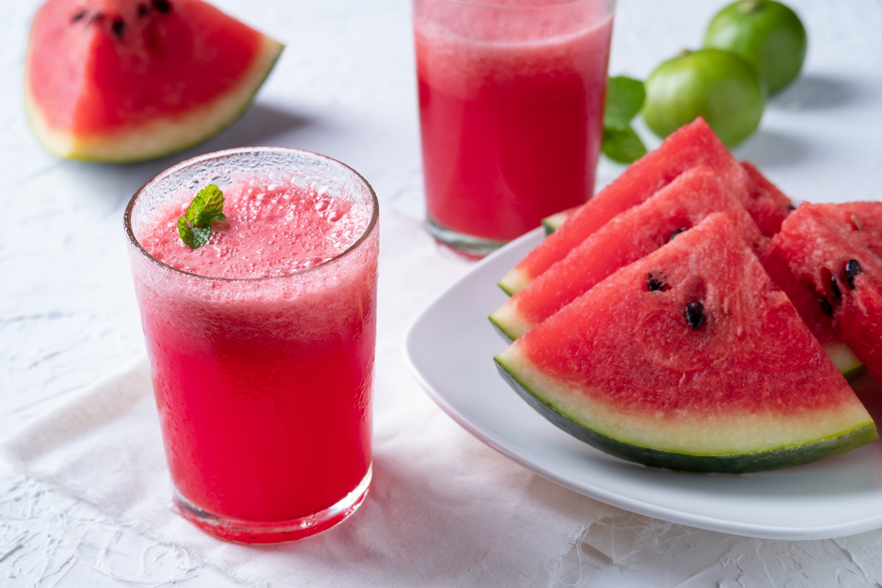 A glass of watermelon juice garnished with fresh mint leaves sitting next to a plate of other watermelon slices.