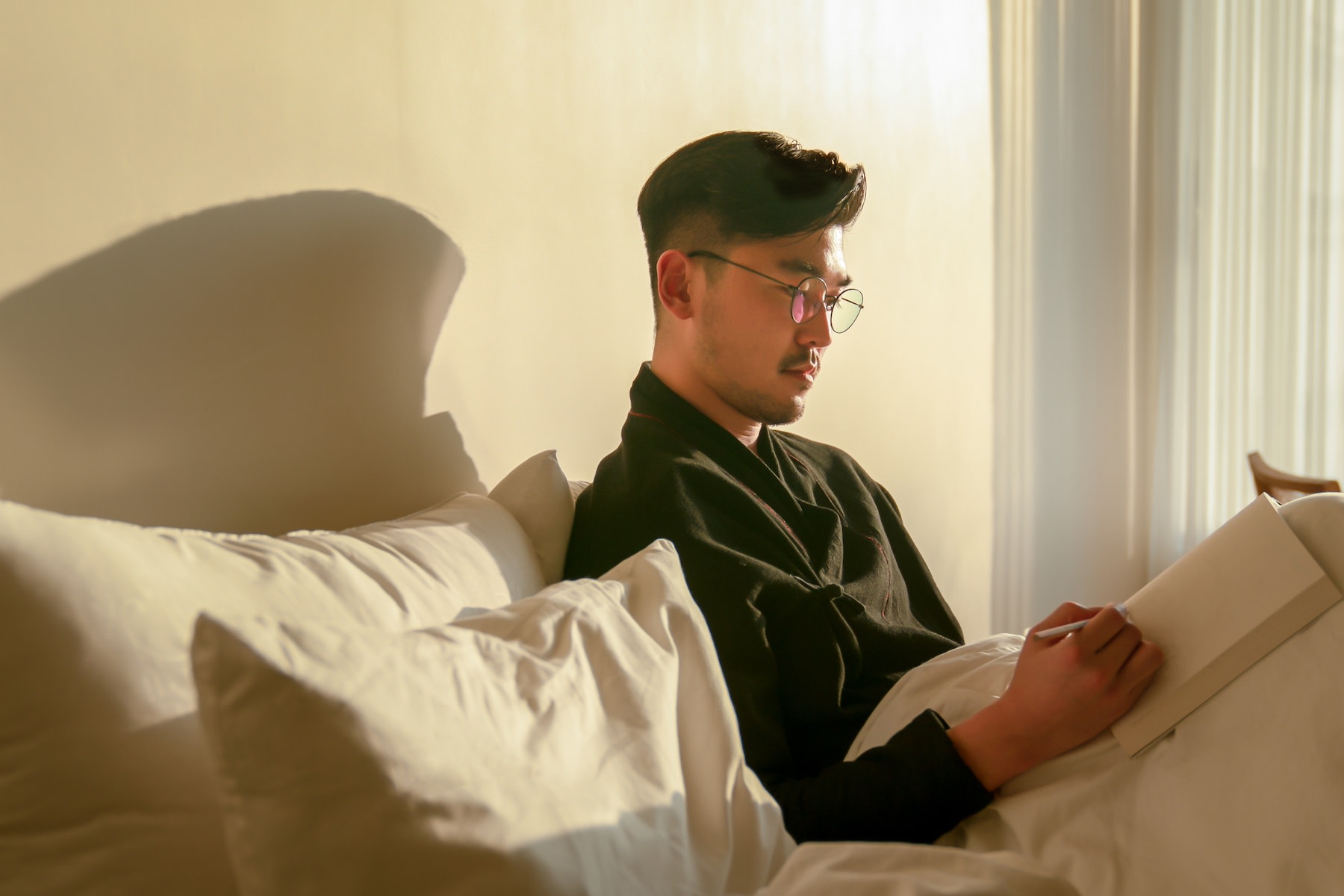 A man sitting up in bed in a sunny room while he journals for mental health.