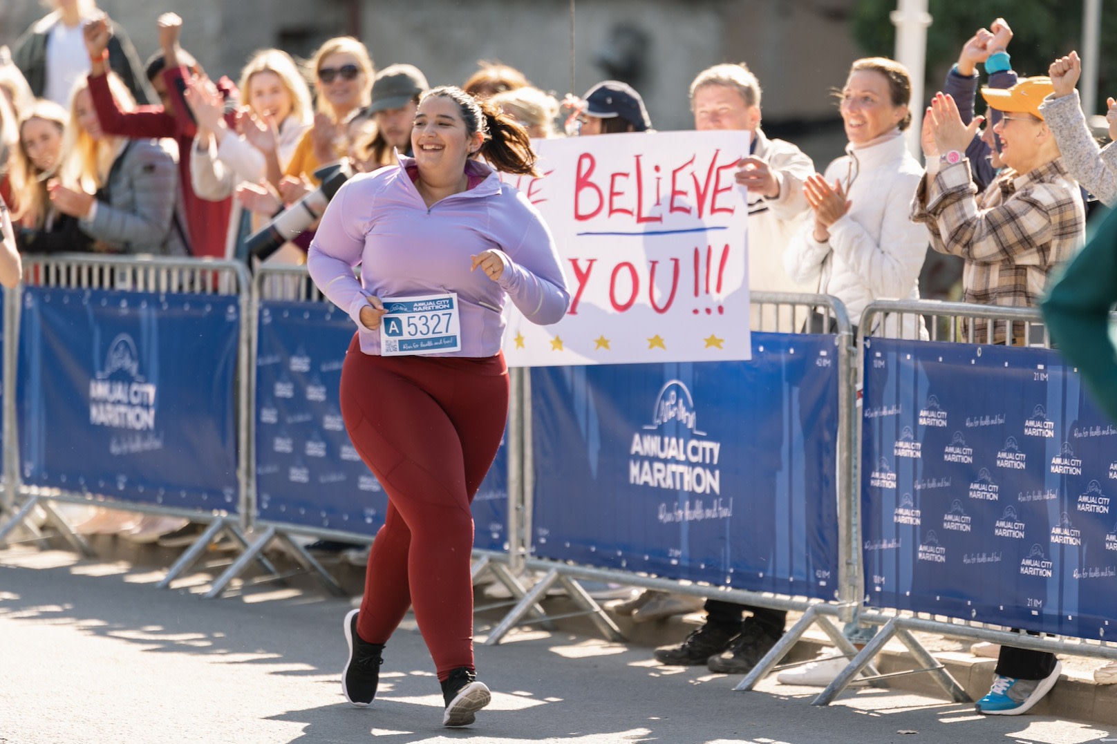 A woman happily running a marathon while a spectator behind her holds up a marathon sign that says "We believe in you!"