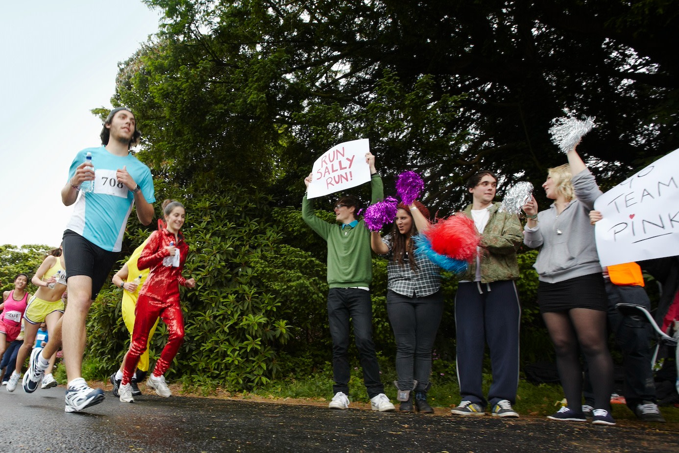 A small crowd of people cheering for runners during a race. A couple of spectators are holding marathon signs that say "Run Sally Run" and "Go Team Pink."