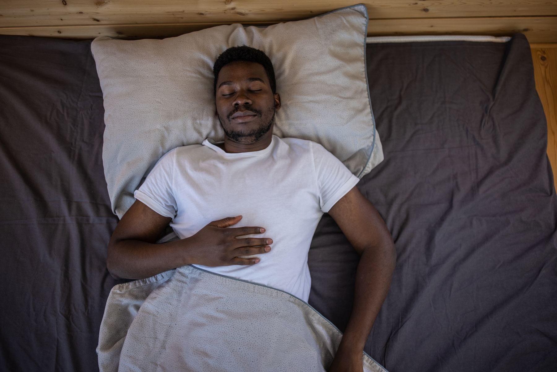 A man practicing breathing exercises for sleep. He's lying on his back in bed, eyes closed, with his right hand on his upper abdomen.