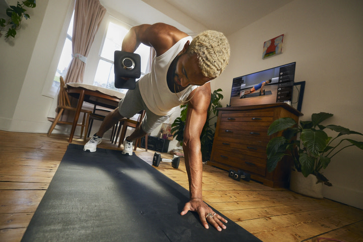 Man weight lifting on workout mat in plank position with Peloton class playing in the background