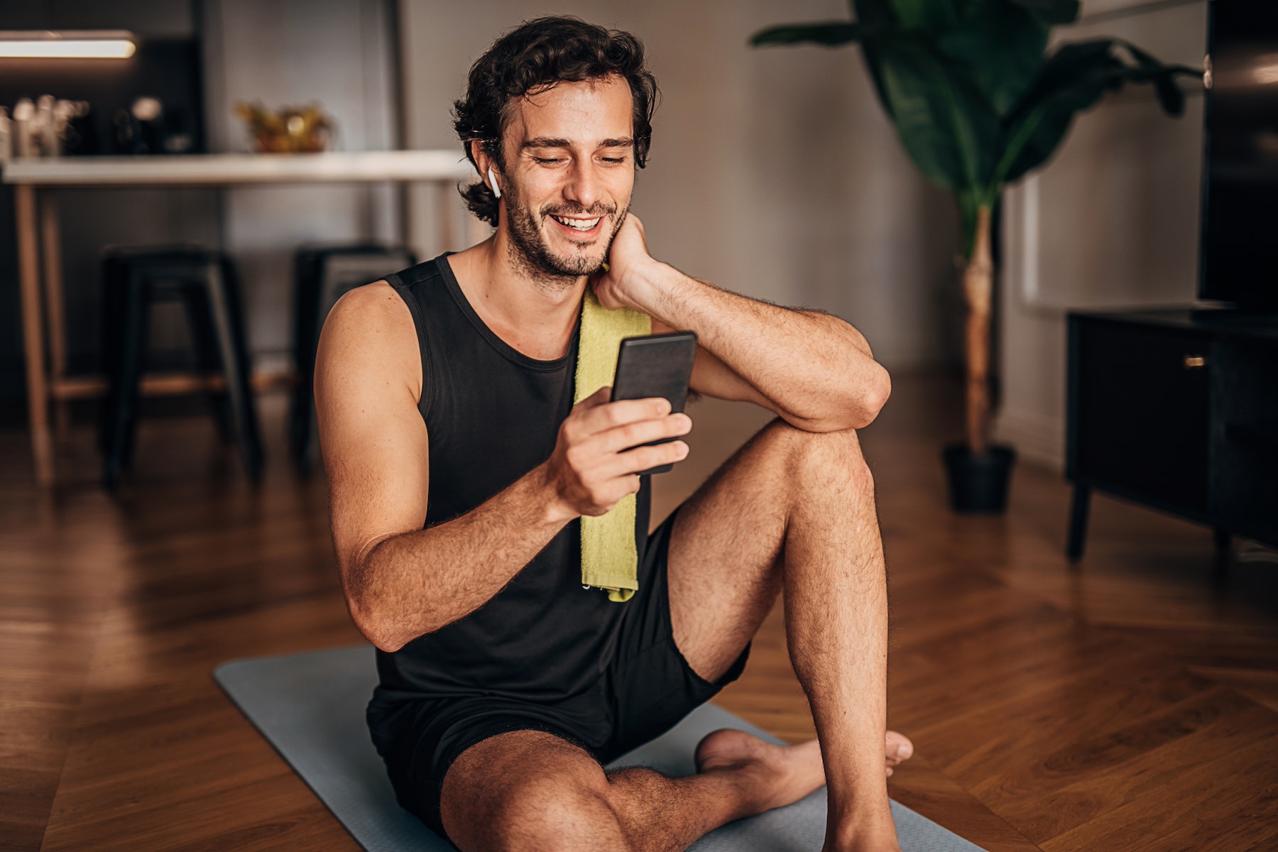 A happy man listening to music on his phone after a workout while sitting on a yoga mat at home. Exercise is a great way to boost your happy hormones.