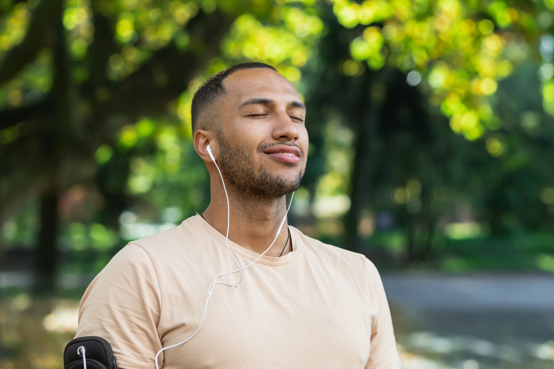 A man practicing mindfulness and meditation. He is closing his eyes and smiling while listening to wired headphones in an outdoor setting.