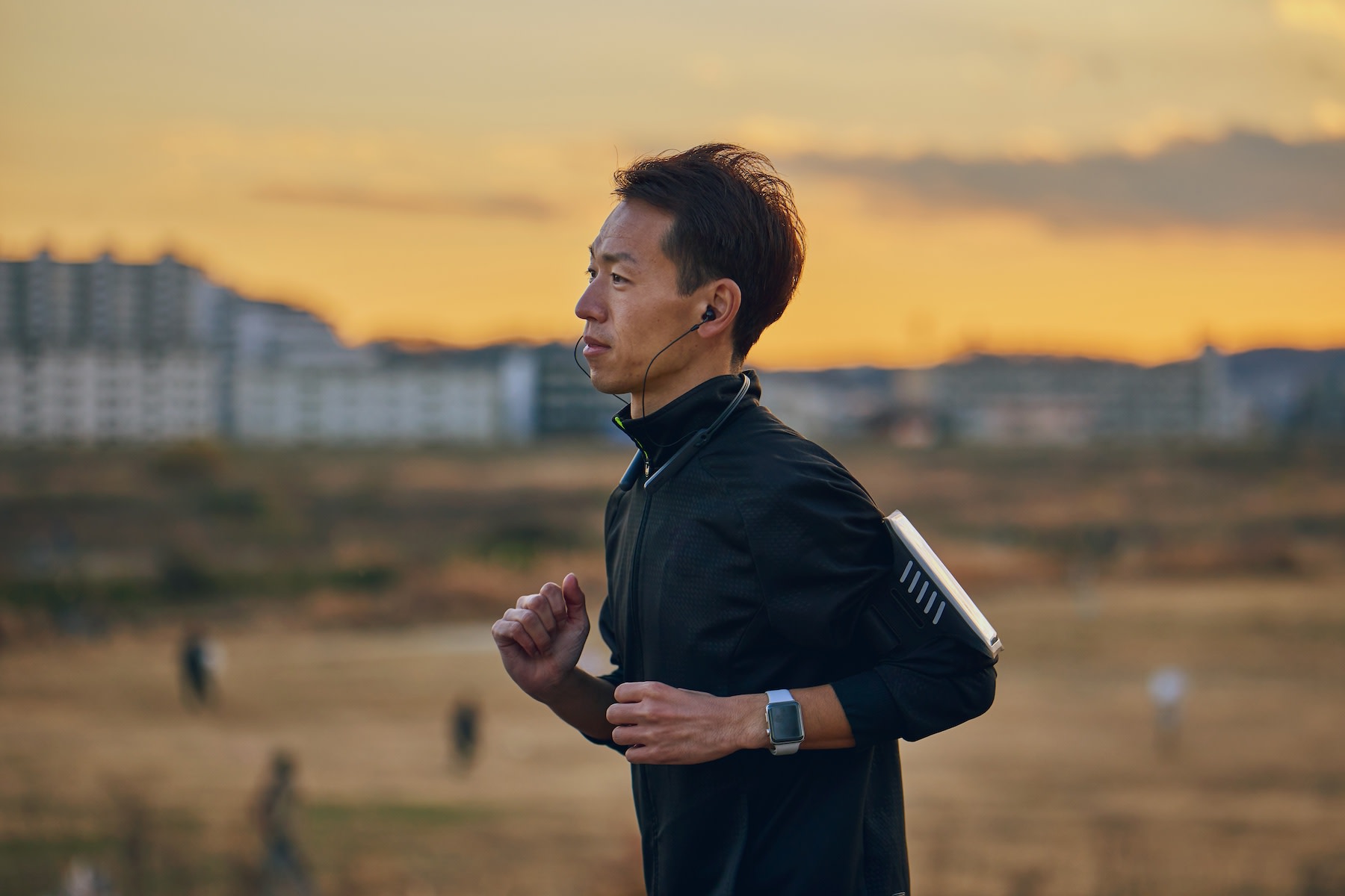 A man going for a jog at dusk. He's listening to headphones and wearing black.