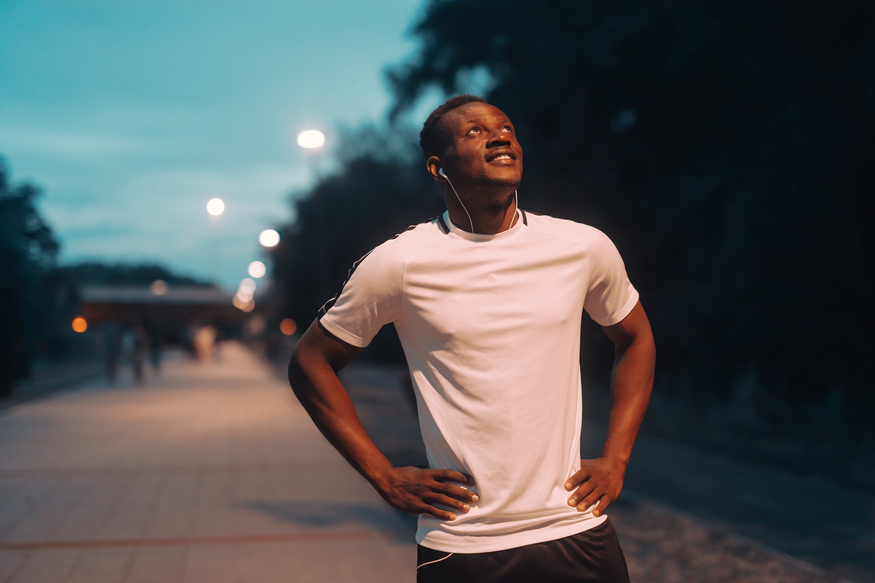 A man recovering from an outdoor walk or jog at night while looking up at the sky and putting his hands on his hips. 